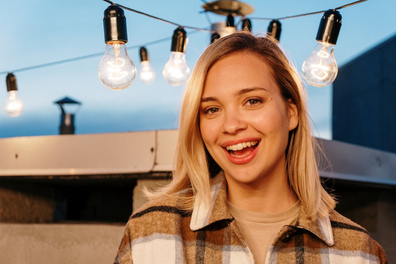 Portrait of a smiling woman on a rooftop with glowing string lights at twilight.